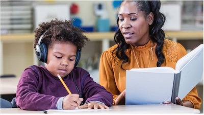 Boy wearing headphones is writing on a desk as his teacher sits beside him and reads out loud