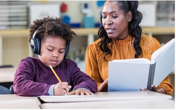 Young black boy wearing headphones writes at a desk while his female teacher dictates to him