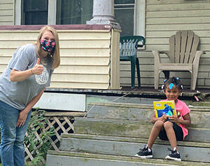A woman with her thumb up, wearing a mask next to a child on porch steps holding a book