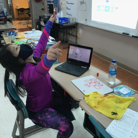 A child at a school desk with their hands up in the air and their head thrown back