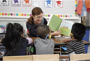 A teacher in front of a group of children pointing at a book.