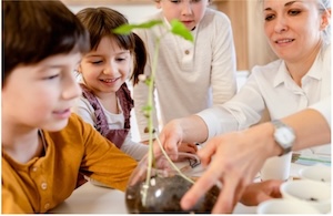Three young students and their teacher observing a seedling plant in a glass test tube