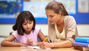 Female teacher sitting next to a girl in a classroom and showing her how to write something in a notebook.