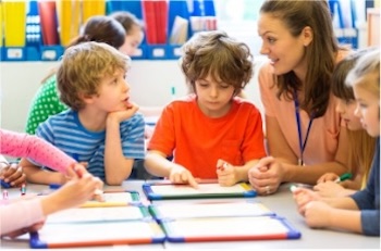 Female teacher and several elementary-aged students seated around a table, working with dry-erase boards