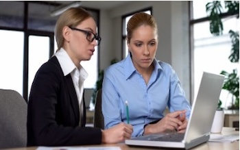 Two women at a laptop computer, one taking notes