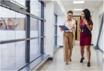 Two women in discussion while walking down a hallway, one holding an open notebook