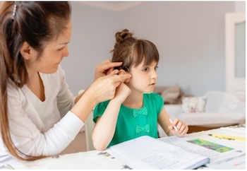 A woman with brown hair helping a young girl with brown hair put in her hearing aid.